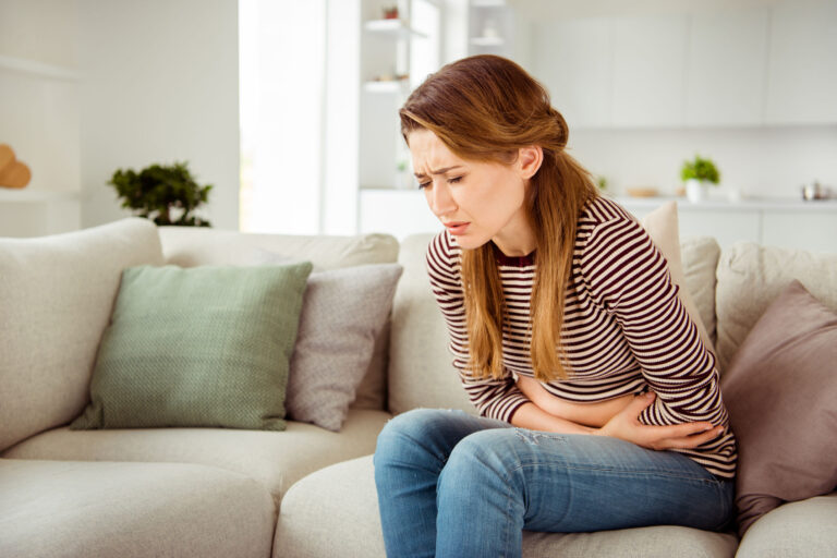 Woman with celiac and ongoing gluten symptoms which may be due to SIBO and IBS but also other things- Image of young woman sitting on couch holding stomach. She is wearing jeans and a sweater and looks very uncomfortable.
