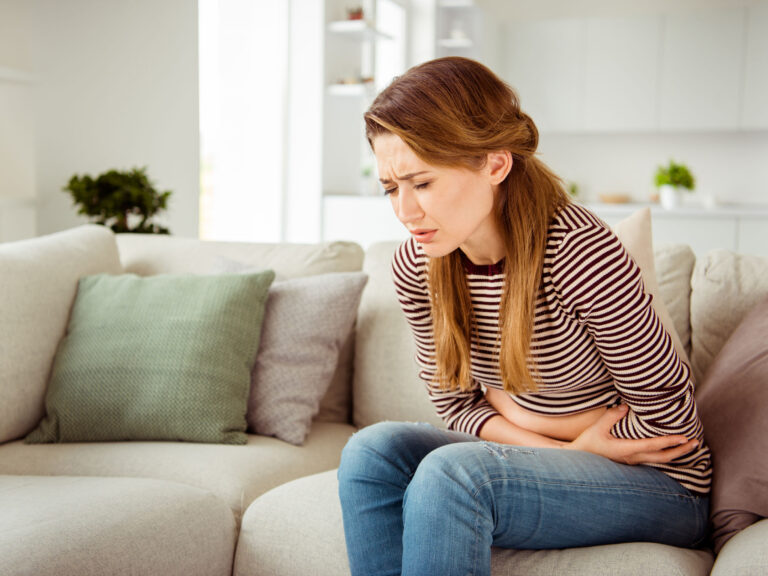 Woman with celiac and ongoing gluten symptoms which may be due to SIBO and IBS but also other things- Image of young woman sitting on couch holding stomach. She is wearing jeans and a sweater and looks very uncomfortable.