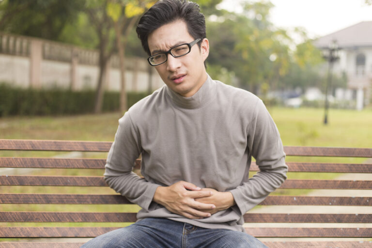 Man with SIBO and IBD sitting on a bench holding his stomach. He is wearing glasses and has a long sleeve t shirt and jeans on.