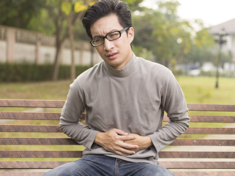 Man with SIBO and IBD sitting on a bench holding his stomach. He is wearing glasses and has a long sleeve t shirt and jeans on.