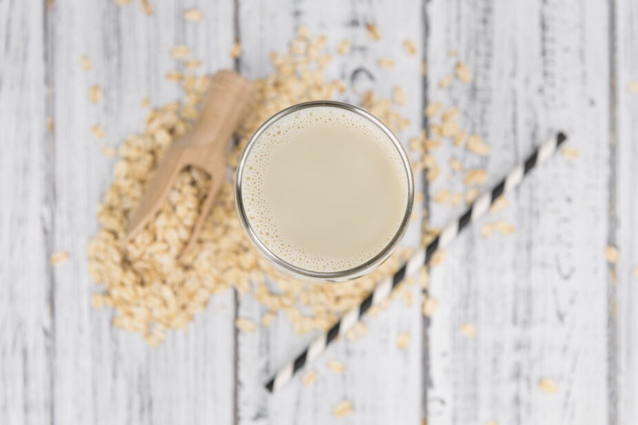 is oat milk gluten free? Image is a glass of oat milk on a table sitting next to a straw and dry oats