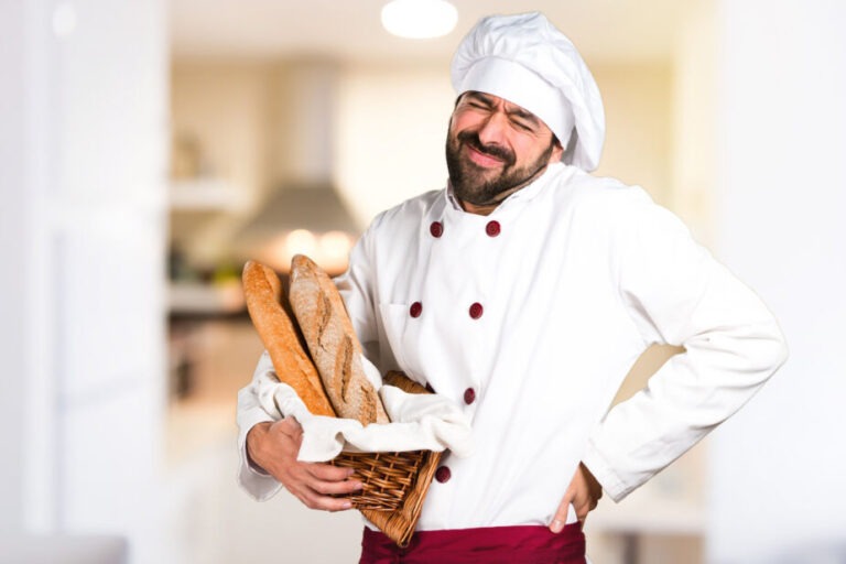 back pain gluten baker in white jacket and hat holding baguettes and his back looking like he is in pain. El dolor de la espalda y gluten un hombre colgando su espalda y llevando pan
