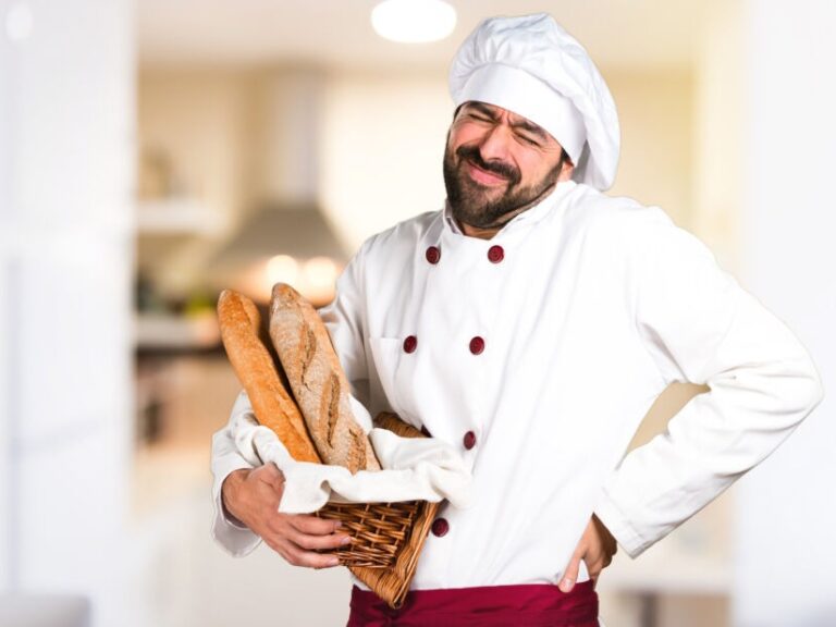 back pain gluten baker in white jacket and hat holding baguettes and his back looking like he is in pain. El dolor de la espalda y gluten un hombre colgando su espalda y llevando pan