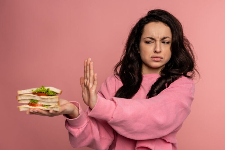 Displeased young dark-haired female showing her food aversion during the studio photo shoot on the pink background. Self-control and nutrition concept arfid in adults is on the rise. arfid symptoms and arfid treatement are explained. Image is of a woman refusing food