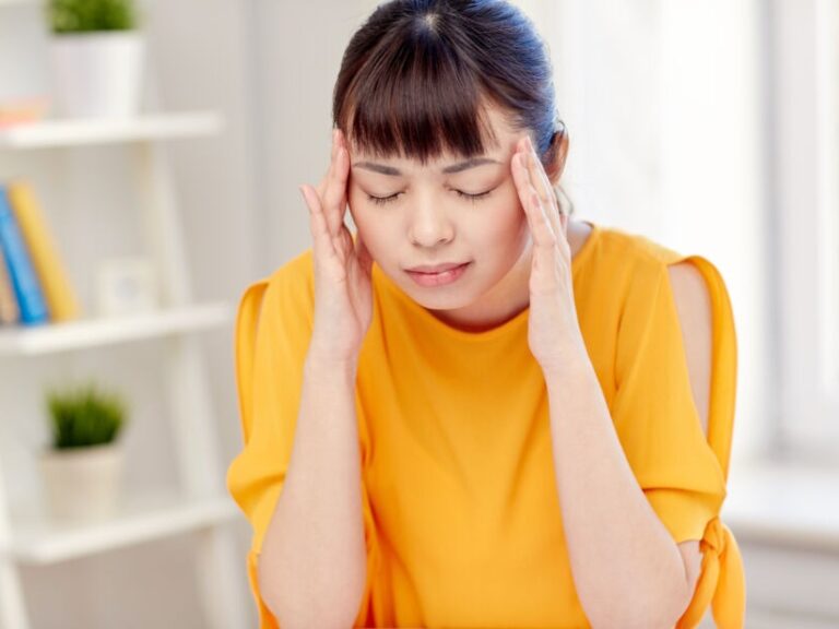 Asian woman in yellow shirt holding her head from a gluten headache. Gluten migraines are common