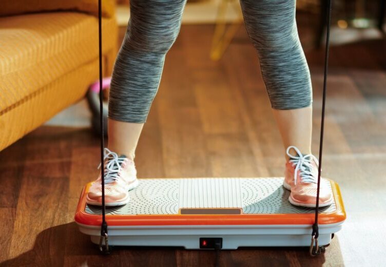 Woman standing on a whole body vibration plate to do exercise and experience whole body vibration therapy and vibration plate benefits. Warable vibration devices have benefits too