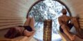 two adults in a barrel sauna with a view window to the forest.