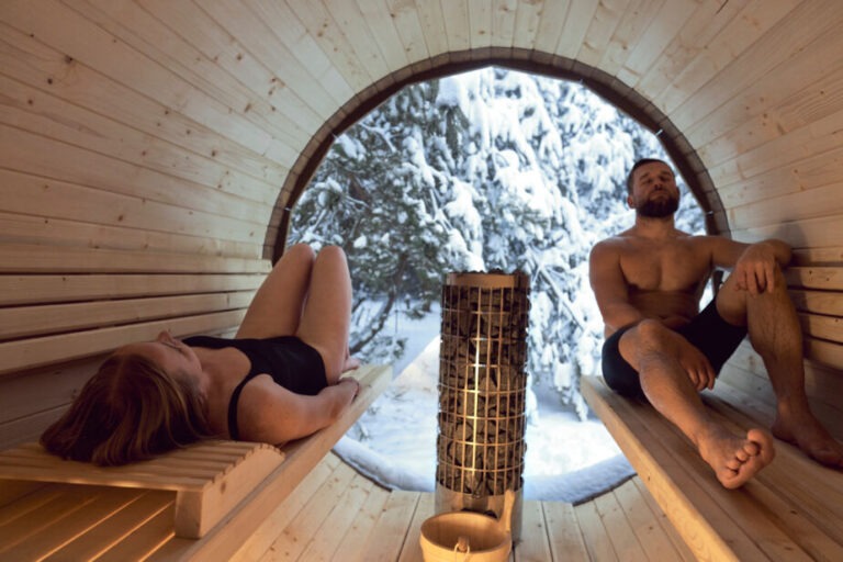 two adults in a barrel sauna with a view window to the forest.